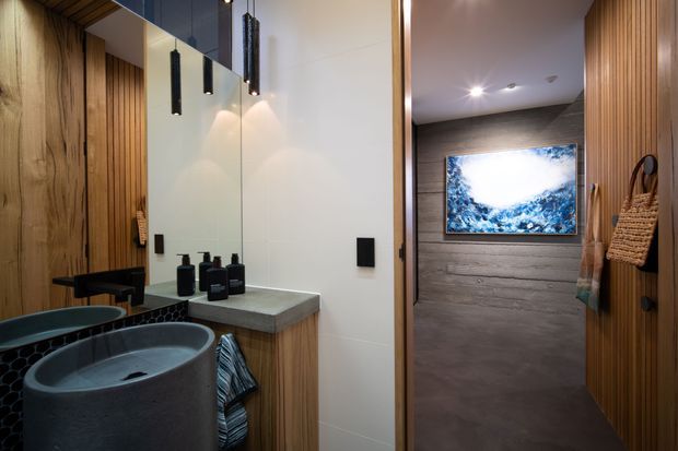 A contemporary bathroom with a dark vessel basin, concrete-look vanity benchtop, timber wall panelling and vertical timber battens. A large mirror reflects the vanity area, while a shower or adjoining wet area is visible through the opening, finished with dark textured wall surfaces and a framed artwork. Black pendant lights and matte black fixtures add contrast.