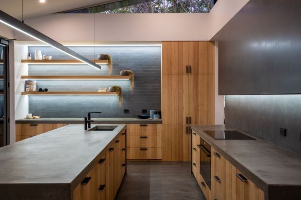 A contemporary kitchen with a large island, timber cabinetry, dark grey concrete-look benchtops, and a charcoal tiled feature wall. Open timber shelves display crockery and small decorative items, while integrated appliances and a cooktop line the perimeter joinery. Linear pendant lighting and concealed shelf lighting illuminate the space, with a narrow clerestory window providing natural light.