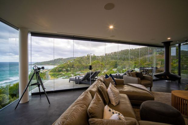 A luxurious contemporary living room with a large curved glazed wall overlooking a coastal hillside and ocean. The room features a deep sectional sofa, armchairs, a dark polished floor, recessed ceiling lighting, a freestanding suspended fireplace, and a telescope positioned by the windows. The expansive glazing creates a strong indoor-outdoor connection and panoramic coastal views.