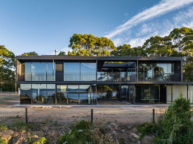 Exterior view of a contemporary two-storey house with a long, rectilinear dark-framed facade, extensive floor-to-ceiling glazing, upper-level balconies, and an open central stair or breezeway. The home sits in a landscaped setting with mature trees and distant water visible through the glazing.