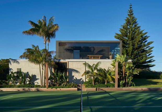Exterior view of a contemporary two-storey residence behind a full-size tennis court. The home combines a pale rendered lower level with a dark, box-like upper volume featuring extensive glazing and a covered balcony. Tropical palms, a tall conifer, landscaped planting, and strong afternoon shadows frame the property.