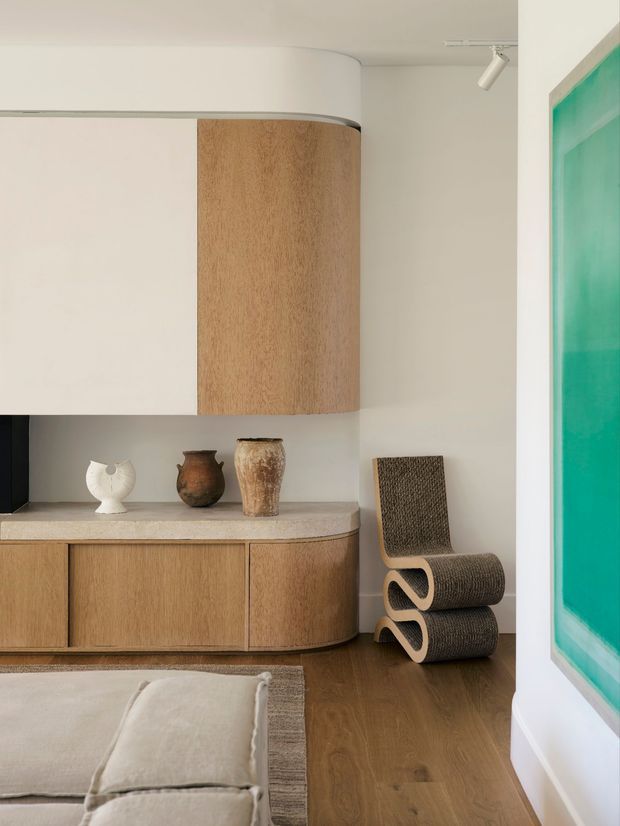 A refined contemporary living room vignette featuring curved built-in timber cabinetry and a matching rounded upper cabinet, a pale stone benchtop, warm timber flooring, and neutral plaster walls. A sculptural woven lounge chair sits beside the cabinetry, with ceramic and sculptural objects displayed on the counter. A large vivid turquoise artwork adds a strong accent at the right edge, while a low neutral sofa and rug are partially visible in the foreground. The space has clean lines, soft curves, warm natural materials, and restrained styling.