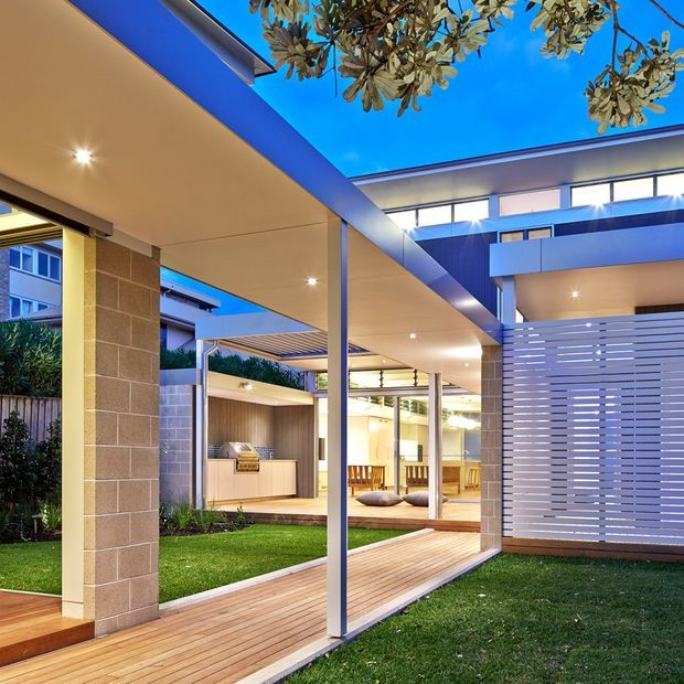 A contemporary residential courtyard viewed from beneath a covered outdoor walkway. The open-plan design features broad roof overhangs, slim steel posts, warm timber decking, pale masonry columns, extensive glazing, and a horizontal slatted privacy screen. A compact outdoor kitchen and dining area are visible beyond the courtyard, with landscaped lawn and mature planting framing the space. The image is photographed at dusk or early evening with interior and recessed exterior lighting creating a warm, inviting atmosphere.