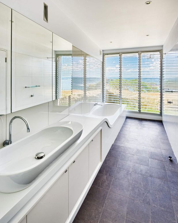 Bright, elongated contemporary bathroom with two sculptural white vessel basins set into a long white vanity, mirrored wall cabinetry, and a freestanding white bathtub positioned beside full-height louvred windows. The windows provide expansive coastal or ocean views and filtered daylight. Dark large-format floor tiles contrast with the predominantly white fixtures, cabinetry, walls, and ceiling. The space has a clean, minimalist, luxurious feel.