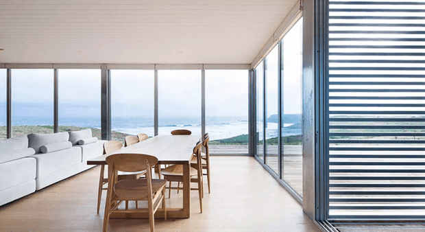Bright, minimalist open-plan living and dining space with pale timber flooring, white upholstered seating, a timber dining table and chairs, and expansive floor-to-ceiling glazing overlooking a coastal landscape. Horizontal exterior screening shades one side of the glazed room.