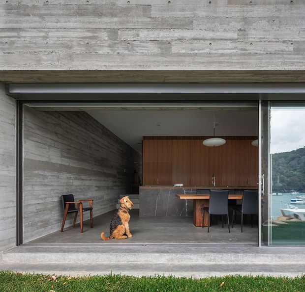 View into a minimalist, open-plan kitchen and dining area through wide sliding glass doors. The space features raw horizontal concrete walls and flooring, dark stone kitchen surfaces, warm timber cabinetry, black dining chairs, and simple pendant lighting. A dog is visible inside, while a waterfront and wooded hillside can be seen through the glazing.