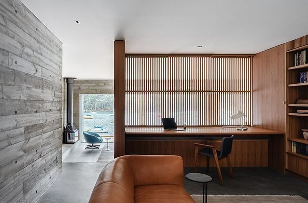 A refined home office or study integrated into an open-plan interior. A long dark timber built-in desk runs beneath a full-height vertical timber slat screen, with a black task chair, desk lamp, shelving, and a tan leather sofa in the foreground. The room features a pale grey concrete-look floor, a white ceiling with recessed lighting, and a dramatic grey timber-clad feature wall. A wood-burning stove and glimpses of an outdoor setting are visible beyond the office.
