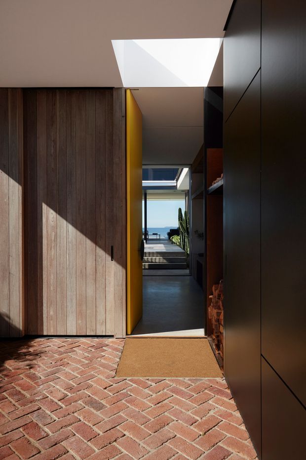 A dramatic architectural view from a brick-paved exterior entry toward a sunlit interior passage and terrace beyond. The composition features a tall vertical timber-clad door or wall on the left, a vivid yellow pivot door, dark smooth wall panels and built-in joinery on the right, exposed brick detailing, and a skylight that casts strong geometric light and shadow. The distant opening reveals blue sky and a coastal-looking horizon.