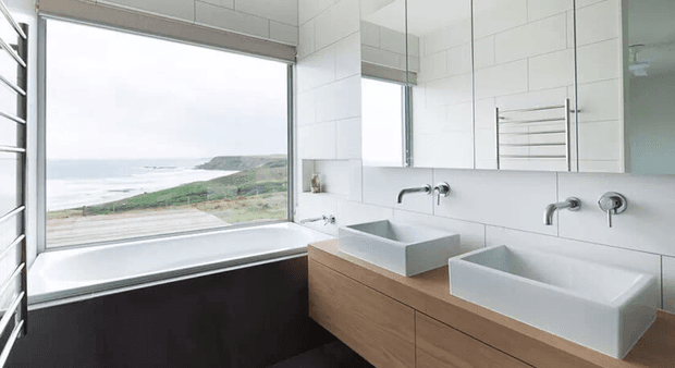 Bright contemporary bathroom with a freestanding-style bathtub set beneath a large picture window overlooking a coastal landscape. A long timber vanity supports two square white vessel basins with wall-mounted chrome taps, while white tiled walls, a large mirror, and heated towel rails complete the clean, minimal fit-out.