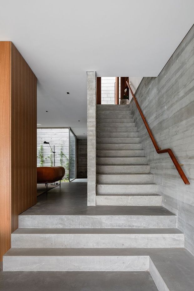 Interior view of a minimalist concrete stairway rising from a broad stepped landing. The space features exposed grey concrete stairs, walls and flooring, a warm timber handrail, vertical timber wall panelling, recessed ceiling lights, and glimpses of a bathtub and planted courtyard beyond.