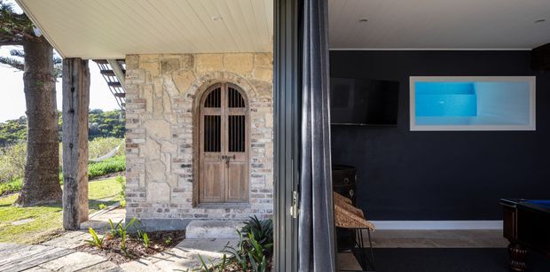 A split view showing a rustic stone-clad entry with an arched timber door and covered verandah on the left, opening into a dark, contemporary media or games room on the right. The interior features charcoal walls, a wall-mounted television, framed artwork, light flooring, and a pool table edge.