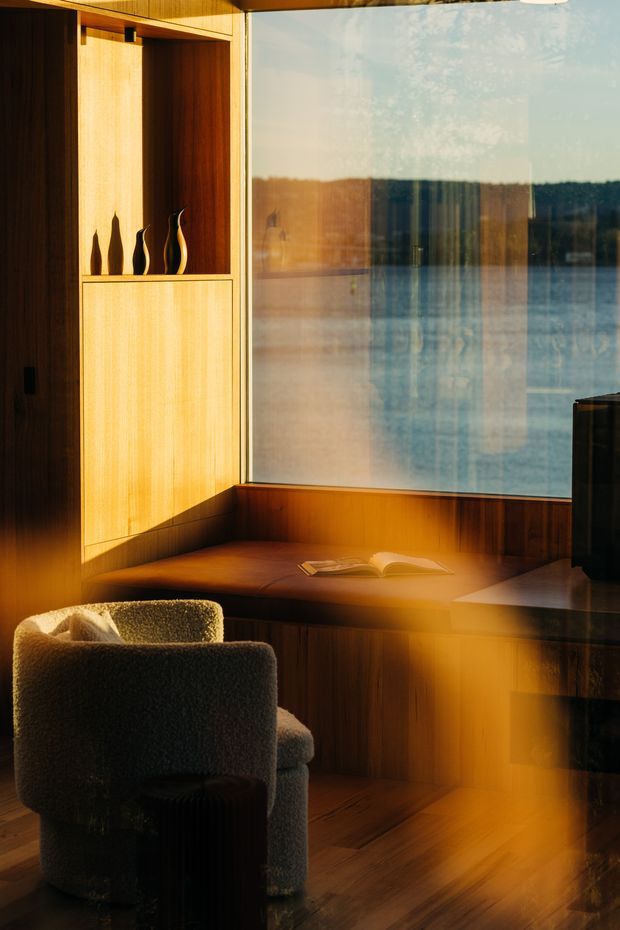 A warm, intimate reading nook beside expansive glazing overlooking a body of water. The space features rich timber built-in cabinetry, a recessed window seat with an open book, a textured upholstered armchair, dark side table, and timber flooring. Strong low-angle sunlight creates dramatic golden reflections across the room and glass.