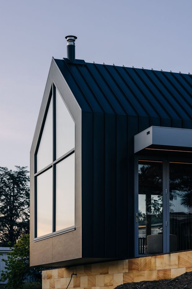 Exterior architectural photograph of a compact contemporary house with a steep gabled roof clad in dark standing-seam metal. The facade combines dark metal cladding with pale fibre-cement or rendered panels, large black-framed glazing, and a projecting entry canopy. The building is elevated on a warm timber-clad base, with a chimney visible above the roofline and trees in the background.