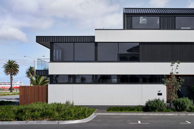 Street-facing view of a contemporary multi-level building with a crisp geometric composition, white rendered wall planes, dark metal cladding and trim, extensive dark-framed glazing, and a projecting upper volume. A small glass balcony, timber privacy screen, landscaped verge, and paved street are visible in front. The building has a flat and low-pitched metal roof profile with strong horizontal bands.