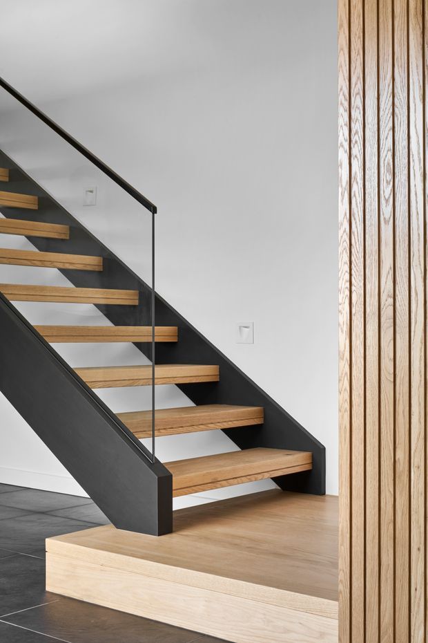 A contemporary interior stairway with open timber treads supported by a dark painted steel or metal structure. A slim glass balustrade with dark handrail runs along the stairs. The stair rises beside a white wall, with a light timber-clad vertical feature wall and light timber flooring/platform; dark tiled flooring is visible at the lower left.