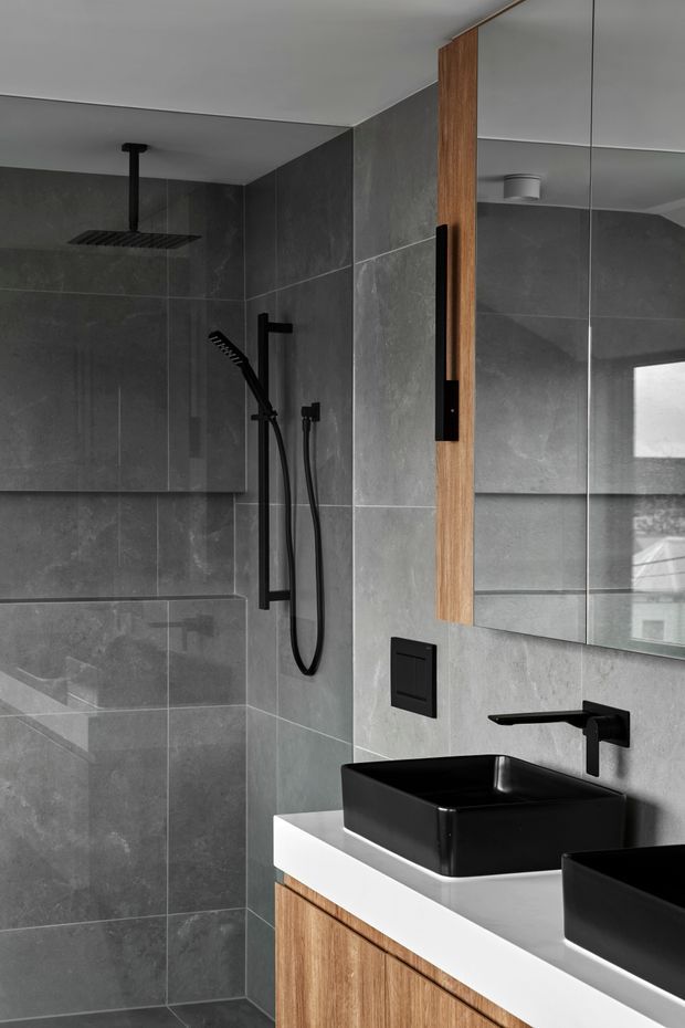 Contemporary bathroom featuring large-format dark grey stone-look wall tiles, a frameless glass shower enclosure, matte black rain and handheld shower fittings, black vessel basins, and a white benchtop over warm timber vanity cabinetry. A full-height mirror and vertical timber detail add contrast.
