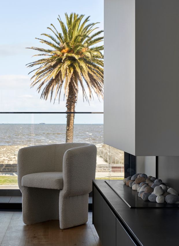 A refined contemporary interior overlooking a waterfront, with full-height glazing framing a palm tree, beach, and water beyond. In the foreground is a sculptural cream bouclé armchair on a light timber floor. A dark, minimalist built-in cabinet runs along the right side, topped with a shallow tray of smooth decorative stones and integrated dark glazing or reflective panels. The space is bright, calm, and coastal in outlook, with strong contrast between the pale upholstery and dark joinery.