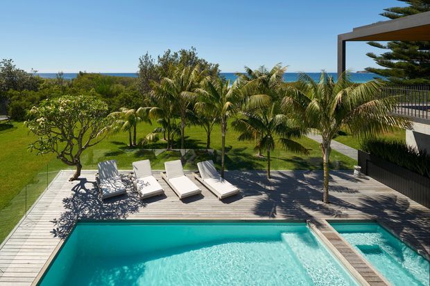 A bright elevated view of a landscaped coastal pool area beside a contemporary multi-level residence. A large turquoise swimming pool with an integrated shallow section occupies the foreground, surrounded by timber decking, white sun loungers, tropical palm trees, manicured lawn, and glass balustrades. The ocean is visible beyond the garden, while a flat-roofed outdoor structure and dark vertical exterior cladding frame the right side.