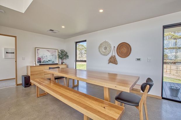 Bright contemporary dining room with a large solid timber dining table and matching bench seating, paired with sculptural timber-and-leather chairs. The room has polished concrete flooring, white walls and ceiling, recessed lighting, broad windows and a glazed door opening to an outdoor area. A timber sideboard and woven wall decorations add warmth.