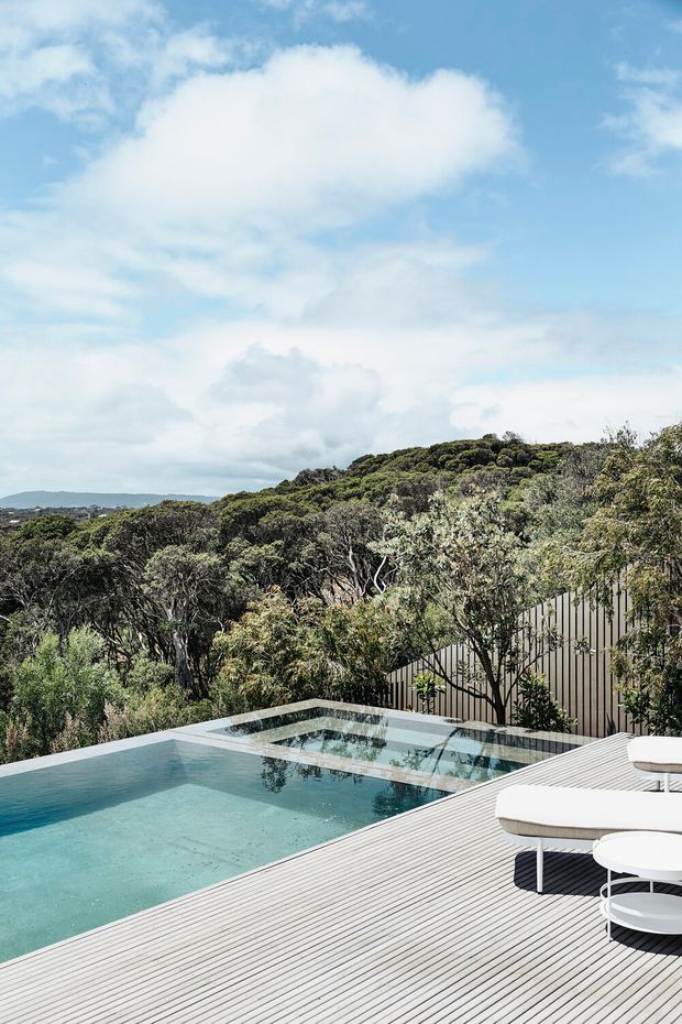 An elevated outdoor pool terrace surrounded by dense native vegetation, with a pale timber-toned deck, turquoise swimming pool, integrated shallow reflective section, and contemporary white outdoor furniture. A vertically ribbed privacy fence lines the edge, while rolling coastal-looking terrain and a bright expansive sky form the background.