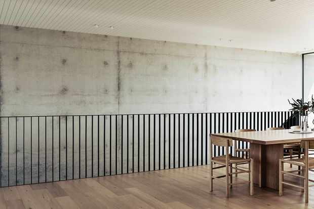 A minimalist dining area on an upper-level landing or mezzanine, featuring a long timber dining table with matching timber chairs, pale timber flooring, a raw concrete feature wall with visible formwork marks, black vertical metal balustrading, and a white linear-panel ceiling. Natural light enters from glazing at the right, with a potted plant and simple tabletop styling.