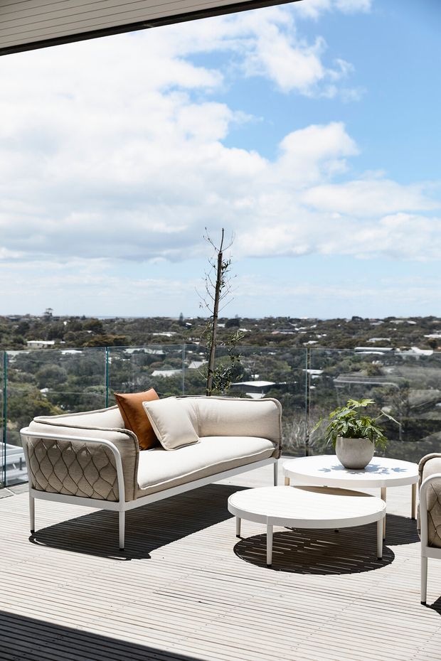 A bright elevated outdoor terrace with pale timber decking and frameless glass balustrades, furnished with a cream upholstered outdoor loveseat, matching armchairs, and layered round white coffee tables. A potted plant provides greenery, while the terrace overlooks a leafy suburban landscape beneath a partly cloudy blue sky. The image has a relaxed, airy contemporary feel.