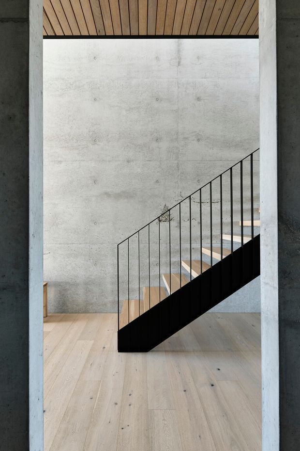 Minimalist interior view of a modern stairway framed by a concrete doorway. The stair has a black solid stringer and slender black vertical balustrade, with light timber treads and pale timber flooring. Exposed concrete walls and a timber-lined ceiling create a restrained, material-focused architectural composition.