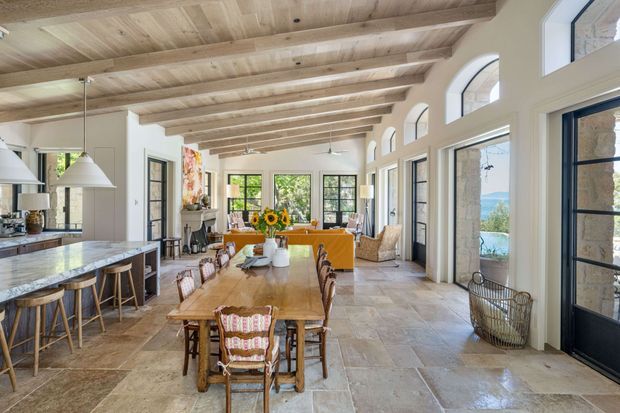A bright, expansive open-plan kitchen, dining, and living area with a long timber dining table, kitchen island, and orange upholstered seating. The room features a high exposed timber-beam ceiling, pale stone-look flooring, white walls, large black-framed windows and doors, and tall arched window openings. Coastal views are visible beyond the glazing, while natural light fills the space.