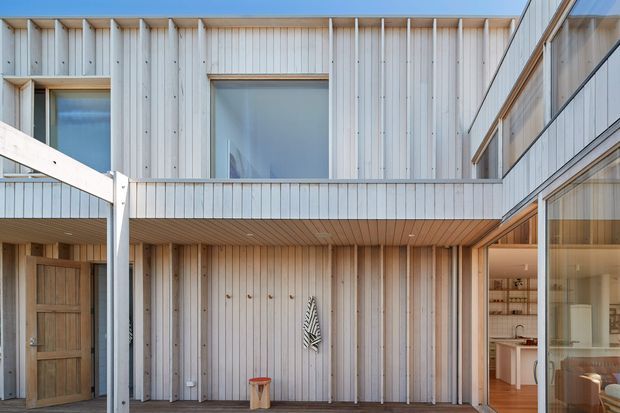 A bright contemporary courtyard view of a two-storey residence. The facade combines pale vertical timber cladding, exposed timber soffits, large glazed openings, and clean-lined balcony and walkway elements. A glazed opening on the right reveals part of a warm timber-lined interior with a kitchen or dining area. The composition emphasises a sheltered central outdoor space and strong indoor–outdoor connection.