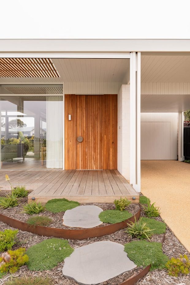 Front exterior view of a contemporary single-storey home, focused on the covered entry porch and large vertical timber front door. The facade combines white vertical cladding, broad white structural posts, timber decking, and glazed openings. A landscaped front garden with curved rust-toned metal edging, gravel mulch, low groundcover, ornamental grasses, and irregular concrete stepping stones leads to the entry. A covered carport is visible to the right, while the left glazing reveals part of the interior living area.