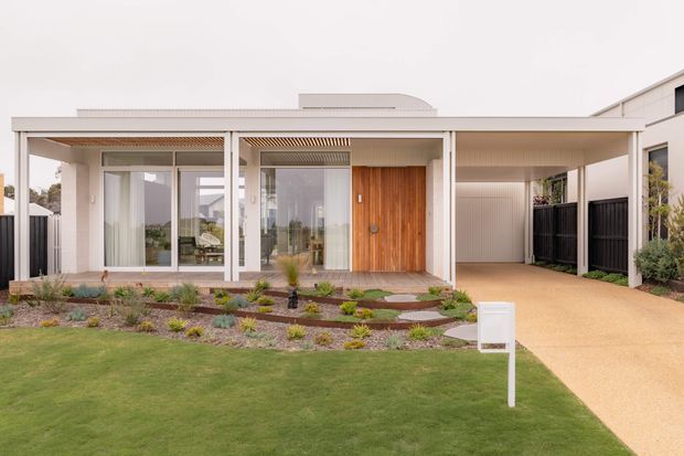 Contemporary single-storey home facade with a broad covered entry veranda, extensive floor-to-ceiling glazing, a warm timber front door, and an integrated carport. The house is set behind a manicured lawn and low, drought-tolerant garden planting, with a pale rendered exterior and crisp white framing.