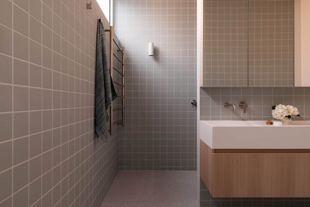 Contemporary bathroom with a walk-in shower and wall-mounted towel rail on the left, and a floating timber vanity with a white integrated basin on the right. The room is clad in small square grey-beige tiles, with warm metallic tapware and soft, understated styling.