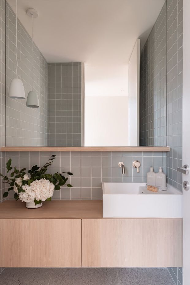 A compact, thoughtfully designed bathroom featuring grey square tiled walls, a large frameless mirror with integrated pendant lights, a white wall-mounted basin, and light timber vanity cabinetry with a matching shelf ledge. A potted floral arrangement adds a natural decorative element, while the terrazzo-style floor and soft neutral palette create a calm contemporary feel.