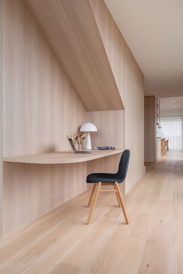 A calm, minimalist interior hallway featuring a built-in pale timber desk and matching wall panelling. A dark upholstered timber-leg chair sits at the desk, which is styled with a white dome lamp, small vase of flowers, and books. The pale timber flooring continues toward a brighter dining or living area in the background. A large angled overhead timber panel creates a distinctive architectural feature above the workspace.