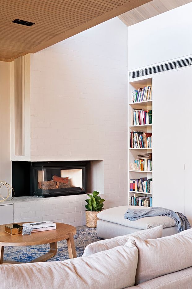 Bright, contemporary living room with a white-painted brick fireplace wall and built-in black firebox, tall integrated open shelving filled with books, and a warm timber slatted ceiling. A pale sectional sofa, round timber coffee table, patterned rug, and potted plant create a relaxed, comfortable seating area. The room has a high ceiling and strong contrast between white surfaces, dark fireplace detailing, and warm timber.