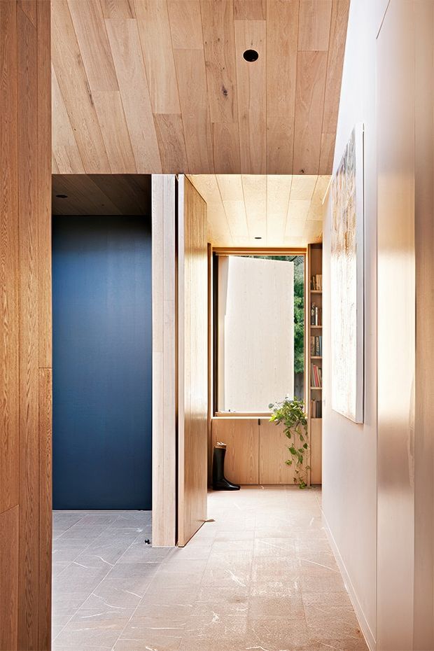 A warm, minimalist interior hallway lined with light timber wall and ceiling panels. The view looks toward a bright room with a large window, built-in shelving, and a potted plant. A dark blue recessed doorway or room opening contrasts with the pale timber and stone-like floor, while a partially open timber door and framed artwork add depth.