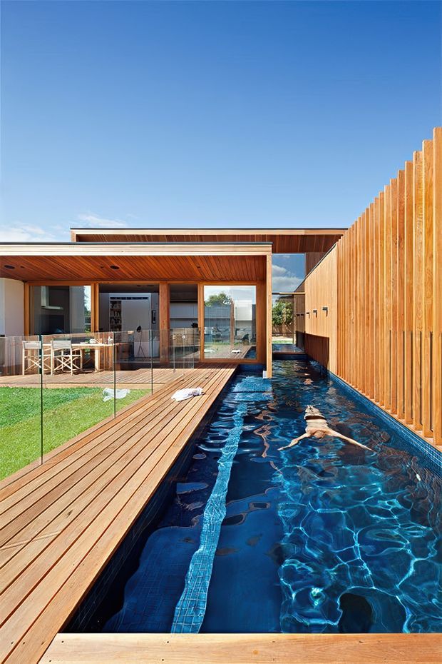 A bright contemporary courtyard-style home viewed from beside a narrow lap pool. The scene features extensive timber decking, a long timber privacy fence, warm timber soffits and posts, floor-to-ceiling glazing, and an open-plan indoor-outdoor living area. The pool dominates the foreground and runs alongside the house, with a clear blue sky above.