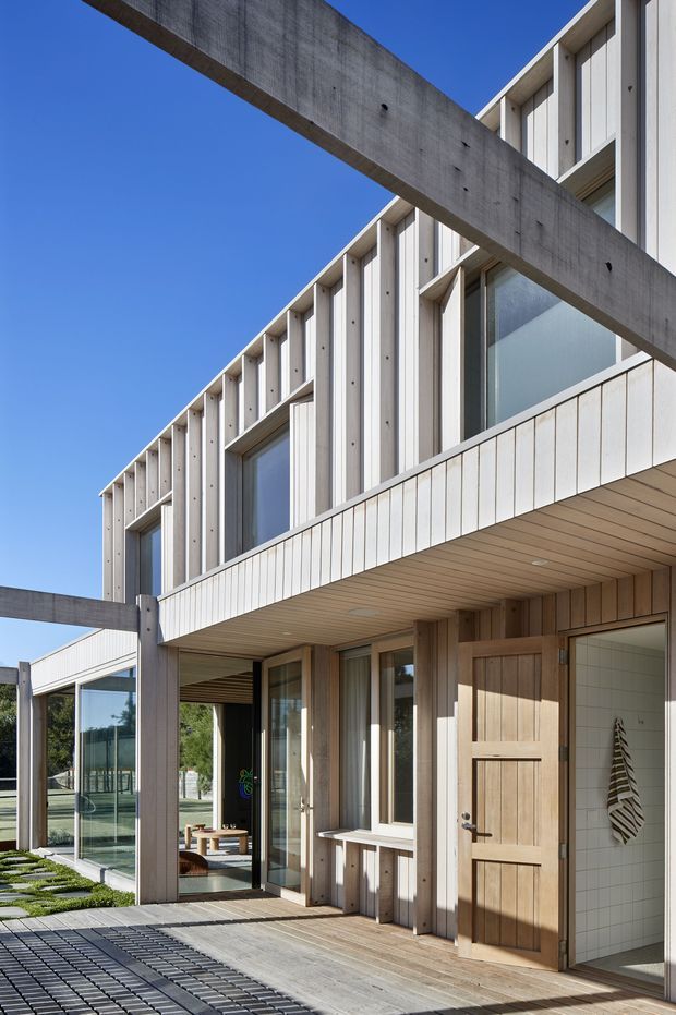 Exterior architectural view of a contemporary multi-level building with pale vertical cladding, extensive glazing, timber detailing, and prominent exposed concrete beams. A covered timber-lined entry and deck lead to an open tiled interior area, while a landscaped courtyard and glass-enclosed zone are visible to the side. The composition emphasises strong horizontal and vertical lines against a clear blue sky.