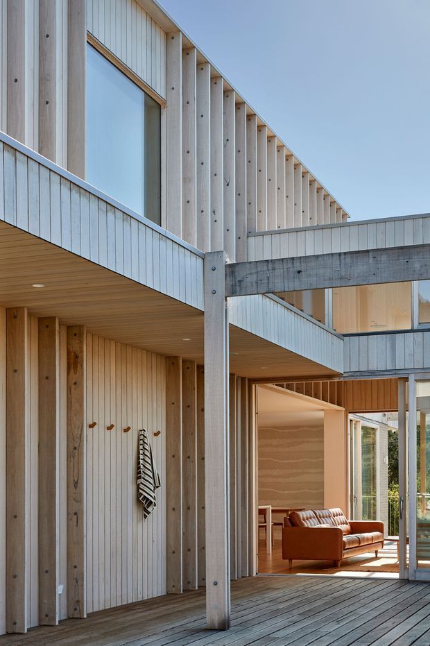 A contemporary multi-level timber-clad home with an open-air covered deck or courtyard. Vertical pale timber siding, exposed structural posts and beams, extensive glazing, and warm timber soffits frame a visible living room furnished with a brown leather sofa. The architecture has strong linear geometry and layered roof and balcony forms.