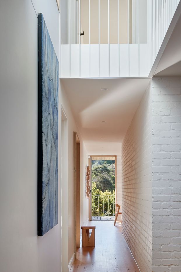 A bright, narrow interior hallway with white-painted brickwork along one wall, smooth white walls and ceiling, and warm timber flooring. A white vertical balustrade overlooks the corridor from an upper-level landing or void. Artwork is mounted on the left wall, while the corridor leads toward a glazed opening and balcony railing with greenery visible outside. A small timber stool and chair add understated furnishing.