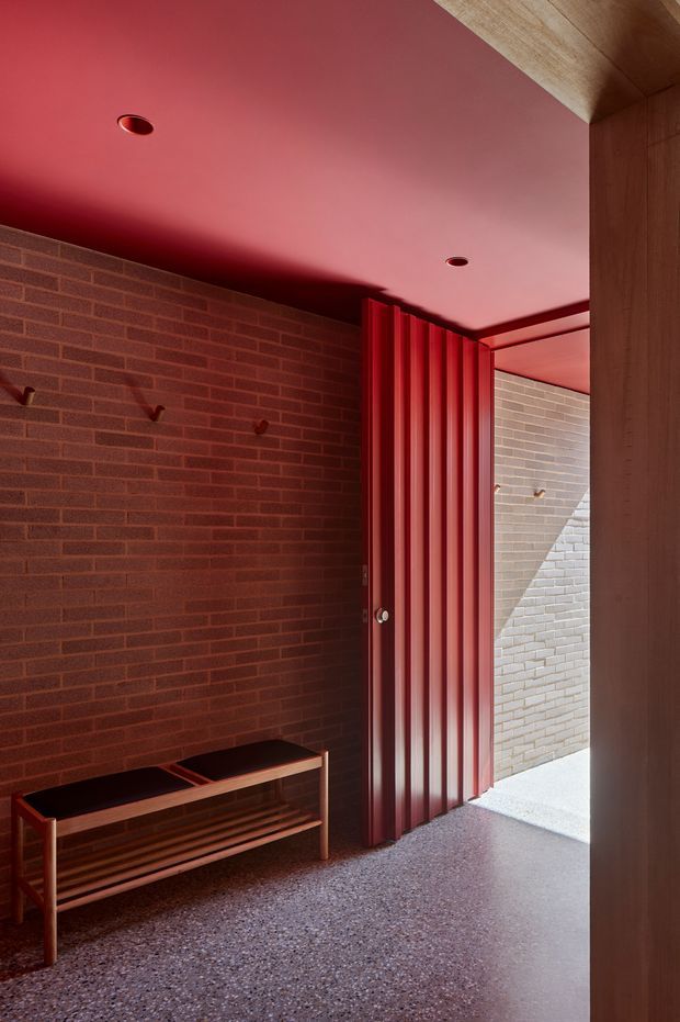 A dramatic architectural entryway with dark red brick walls, a deep red painted ceiling, and a vertically ribbed red metal door opening to a small brick courtyard. A simple timber and dark-top bench with lower slatted shelving sits against the wall, while a timber-lined beam or soffit frames the upper right. The floor has a coarse terrazzo-like aggregate finish, and recessed ceiling lights provide minimal illumination.