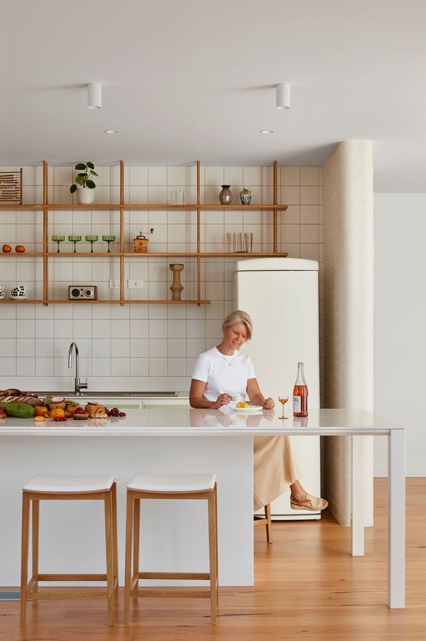Bright contemporary kitchen with a large white island and integrated seating, open timber-framed shelving against a white square-tiled wall, a freestanding white refrigerator, timber flooring, and a curved tiled column. The space has warm wood detailing, minimal white surfaces, and abundant natural light.