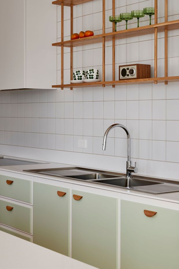 A bright, retro-inspired kitchen focused on a double stainless-steel sink and curved chrome tap. Pale sage-green flat-front cabinetry with warm timber handles sits below a white benchtop and white square-tile splashback. Open timber shelving displays green glassware, ceramics, fruit and a vintage-style radio, adding mid-century character.