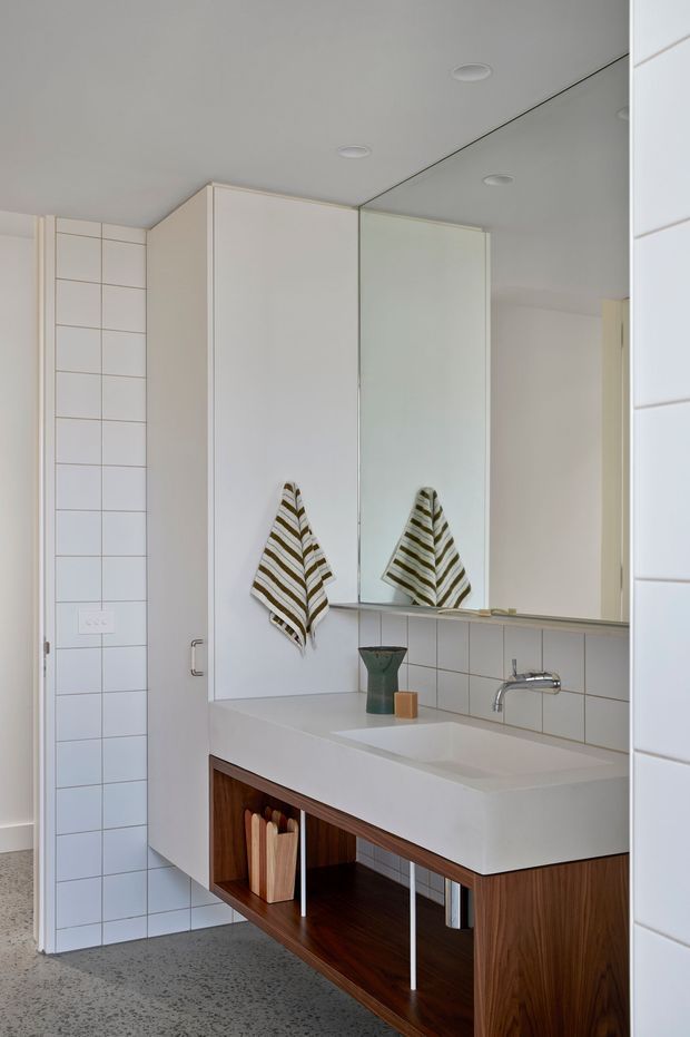 A bright contemporary bathroom featuring a long white integrated vanity basin, dark timber open cabinetry, white rectangular wall tiles, a large mirrored cabinet, and minimal chrome tapware. A striped hand towel and small accessories add subtle warmth. The bathroom has a clean, compact layout with recessed ceiling downlights and a terrazzo-like grey floor.