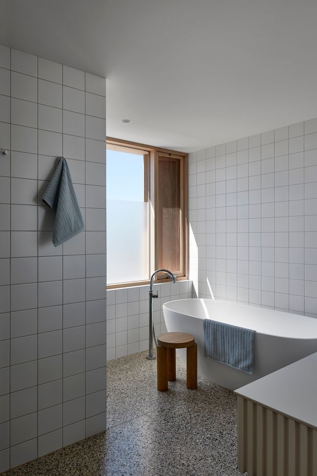 Bright, minimalist bathroom with full-height white square wall tiling, a freestanding white bathtub, timber-framed window and timber panel, chrome floor-mounted bath tap, and a terrazzo floor. Soft blue-grey towels and a small timber stool add subtle contrast. Natural light enters through the window, creating a calm and airy atmosphere.