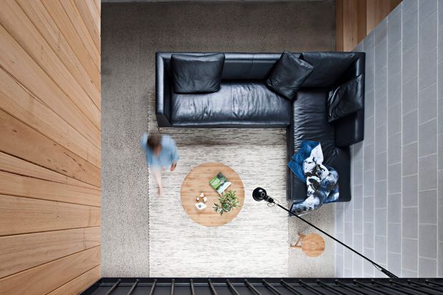 Top-down view of a compact contemporary living room with a black leather L-shaped sectional arranged on a pale textured rug around a round timber coffee table. Warm vertical timber wall panelling lines the left side, while large grey rectangular wall tiles or concrete-look cladding form the right wall. Dark-framed glazing or a balustrade appears along the lower edge, and a blurred person is present near the seating area.