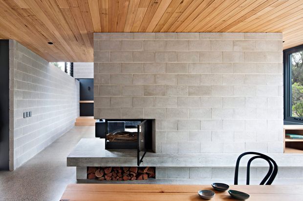 Interior view of a contemporary open-plan living and dining space with a central exposed concrete-block fireplace wall and integrated firewood storage. A long concrete hearth projects into the room, while warm timber boards line the ceiling and dining table. Black-framed glazing and built-in shelving are visible at the right, with a hallway and steps extending behind the fireplace wall.