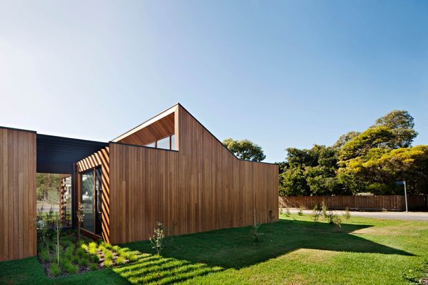 Exterior view of a contemporary single-storey house composed of angular timber-clad volumes, including a prominent gabled wing with a high glazed opening and a darker connecting section. The building sits within a landscaped suburban garden with lawn, planted borders, fencing, and mature trees.
