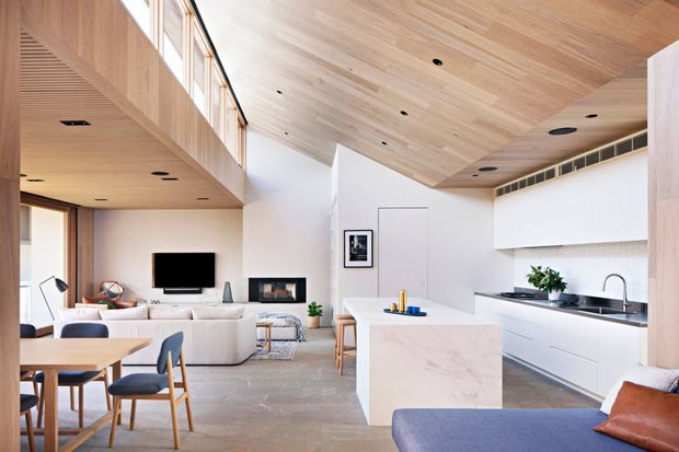 Open-plan contemporary interior combining a kitchen, dining area, and living room. The space features a sculptural white kitchen island with a pale stone benchtop, white handleless cabinetry, integrated appliances, and a stainless-steel sink. Light timber lining spans the ceiling and upper architectural surfaces, with clerestory glazing bringing in abundant daylight. A dining table and upholstered seating occupy the foreground, while a sofa, television, fireplace, and built-in shelving define the living area in the background.