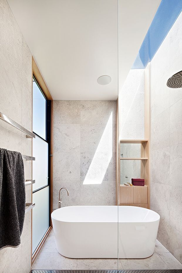 Bright contemporary bathroom featuring a freestanding white bathtub, large pale stone-look wall and floor tiles, a frameless glass shower enclosure, timber-framed window, built-in timber shelving and vanity joinery, and a wall-mounted rainfall shower. Strong natural light creates a clean, airy atmosphere.